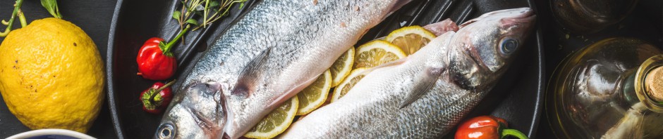Ingredients for cookig healthy fish dinner. Raw uncooked seabass fish with rice, lemon, herbs and spices on black grilling iron pan over dark background, top view, horizontal composition